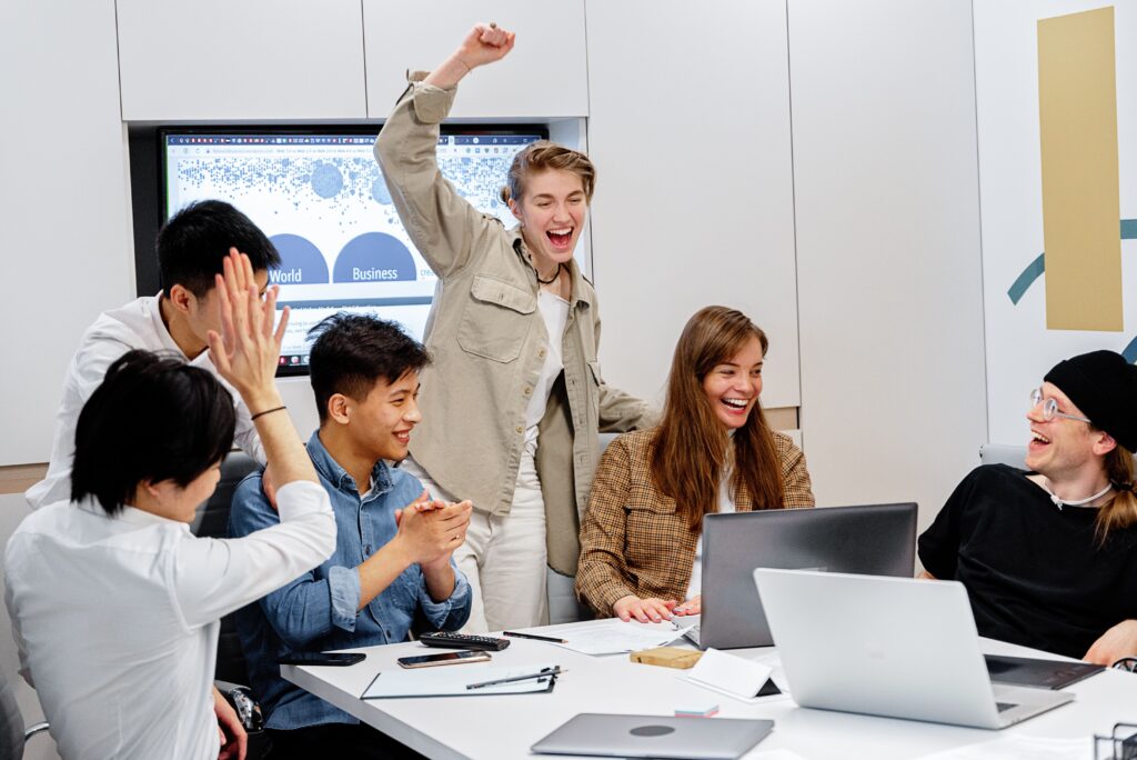 A diverse team enthusiastically celebrating success around laptops during an office meeting.